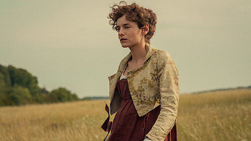 Woman in historical dress standing in a field with tall grass, trees in the background, and a cloudy sky, creating a calm, pastoral setting.