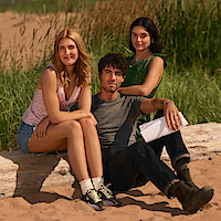 Three young adults sitting on a log at a sandy outdoor area with tall grass, dressed in casual summer clothes; one holding papers, all looking at the camera and smiling, suggesting a relaxed, sunny environment.