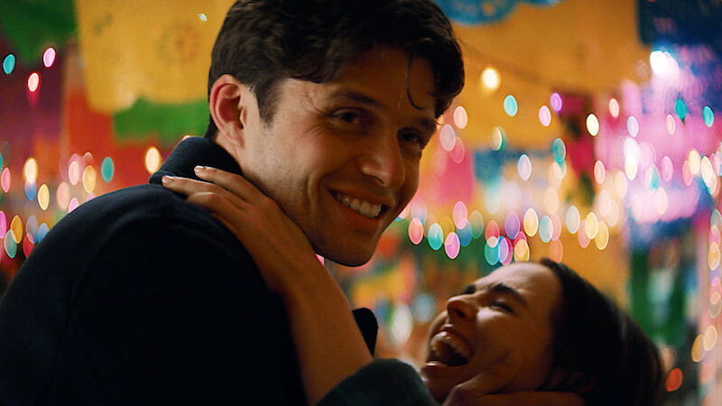 A smiling man embraces a laughing woman at a festive indoor party decorated with colorful lights and banners, creating a joyful, lively atmosphere.