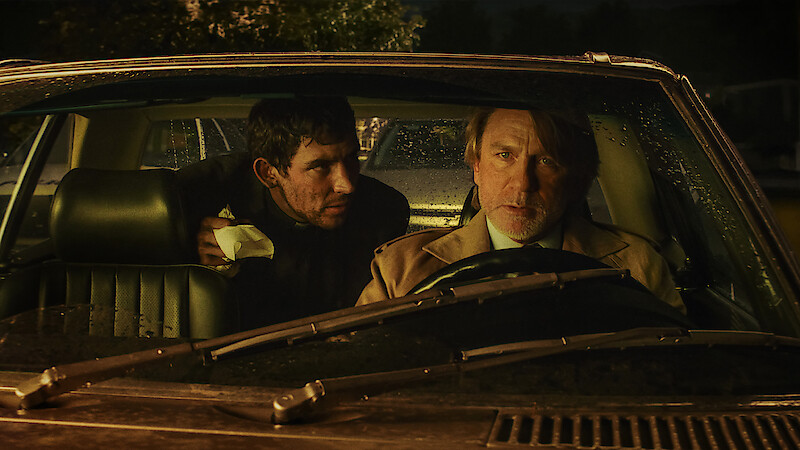 Two men sit in a car at night, illuminated by dim streetlights and rain on the windshield. The driver looks concerned while the passenger leans in with urgency, creating a tense and suspenseful atmosphere.