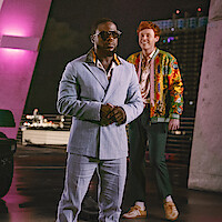 Two men standing outdoors at night, one in a light blue suit and sunglasses, the other in a colorful jacket, with city lights and modern architecture in the background, under pink and purple lighting.