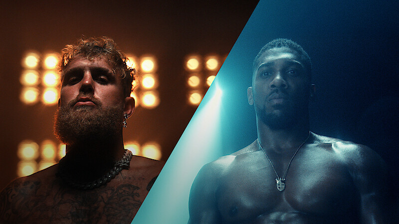 Two muscular men stand shirtless under dramatic lighting, one in warm orange light with stadium lights behind him, the other in cool blue light, both serious and intense, creating a powerful, competitive mood.