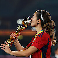 Female soccer player in red jersey kissing a gold trophy on a stadium field, wearing a medal and headband, celebrating victory with blurred stadium lights in the background.