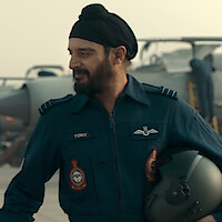A pilot in a blue uniform and turban stands on an airbase holding a helmet, with military aircraft and crew visible in the background.
