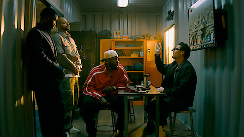 Four men in a dimly lit storage room; three stand while one in glasses gestures, sitting at a table. Shelves, jackets, and a wall map add to the gritty, informal setting.