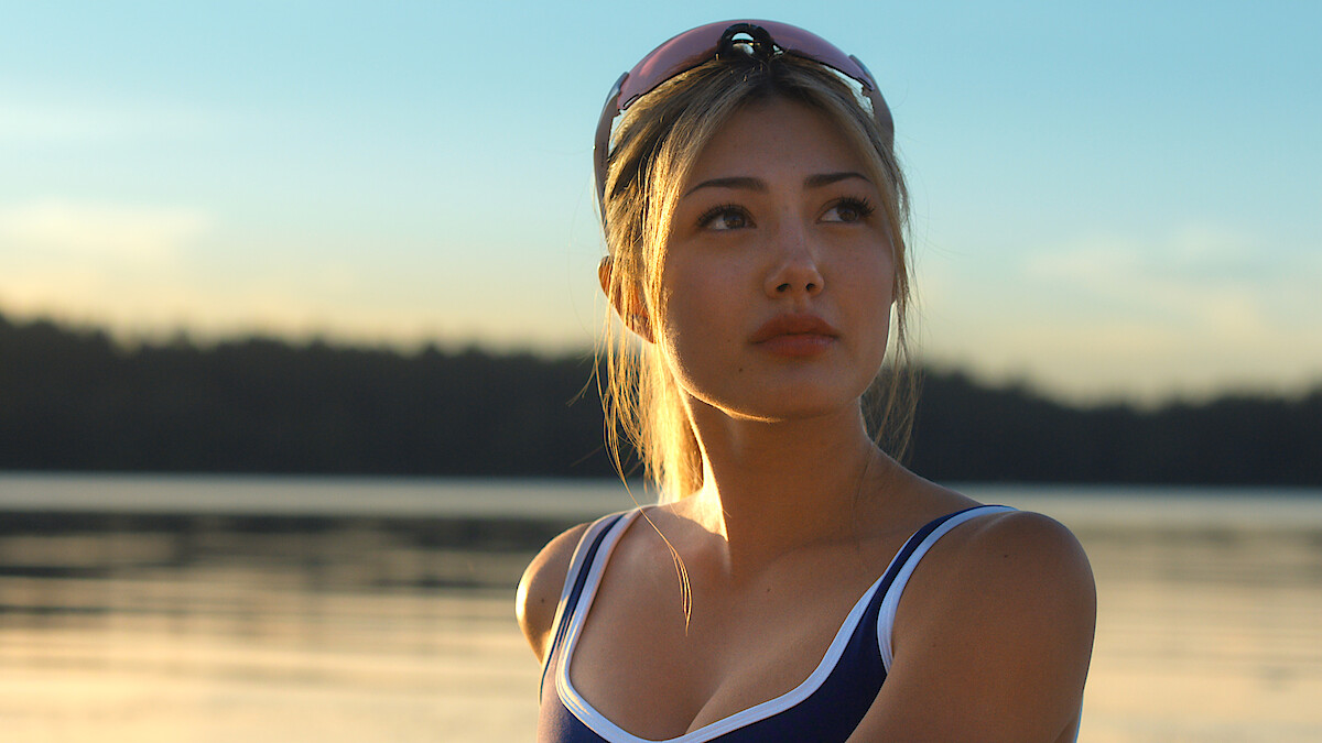 Woman in athletic swimwear with goggles on head stands by a calm lake during sunset, surrounded by serene water and trees in the background, illuminated by soft, natural light.