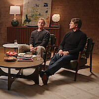 Two men sit in armchairs in a cozy living room with a brick wall, abstract art, and lamps behind them. In front, a coffee table holds books, snacks, and glasses of water, creating a relaxed, conversational setting.