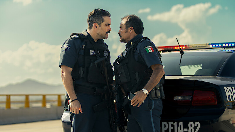 Two uniformed police officers stand face-to-face in a tense confrontation beside a police car with flashing lights on a sunny day, with mountains and a blue sky in the background.