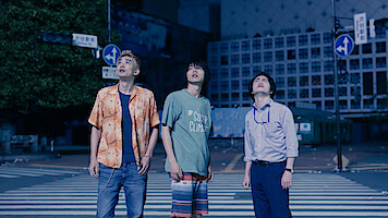 Three young men stand on a city crosswalk at night, looking up with curious or concerned expressions. The urban street is quiet and dimly lit, surrounded by buildings and street signs.