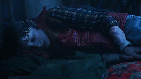 A young boy in a plaid shirt and red vest lies curled up on his side in a dimly lit setting, appearing wet, alone, and cold.