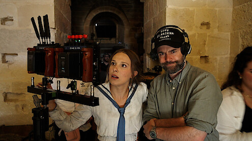 A woman in costume and a man with headphones stand by a camera rig in a stone hallway on a film set, reviewing footage together.