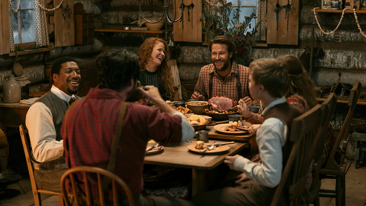 Six people in rustic clothing share a meal and laugh together inside a cozy log cabin, surrounded by wooden furniture, dim lighting, and a festive, warm atmosphere.