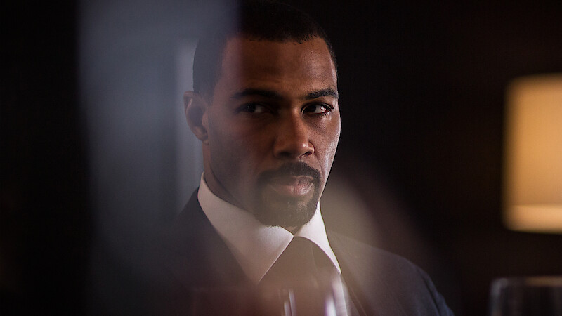 A man in a suit sits in a dimly lit room with a serious expression, blurred glass and a lamp in the background, creating a dramatic, tense atmosphere.