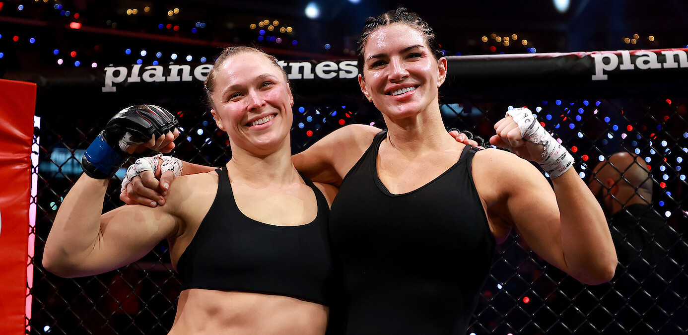 Two female MMA fighters in black sportswear stand arm-in-arm and smiling inside a caged arena after a match, with bright lights and cheering audience in the background.