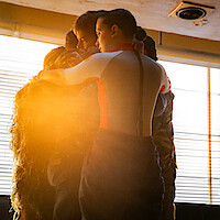 A small group huddles in a tight embrace, backlit by warm sunlight streaming through large window blinds.