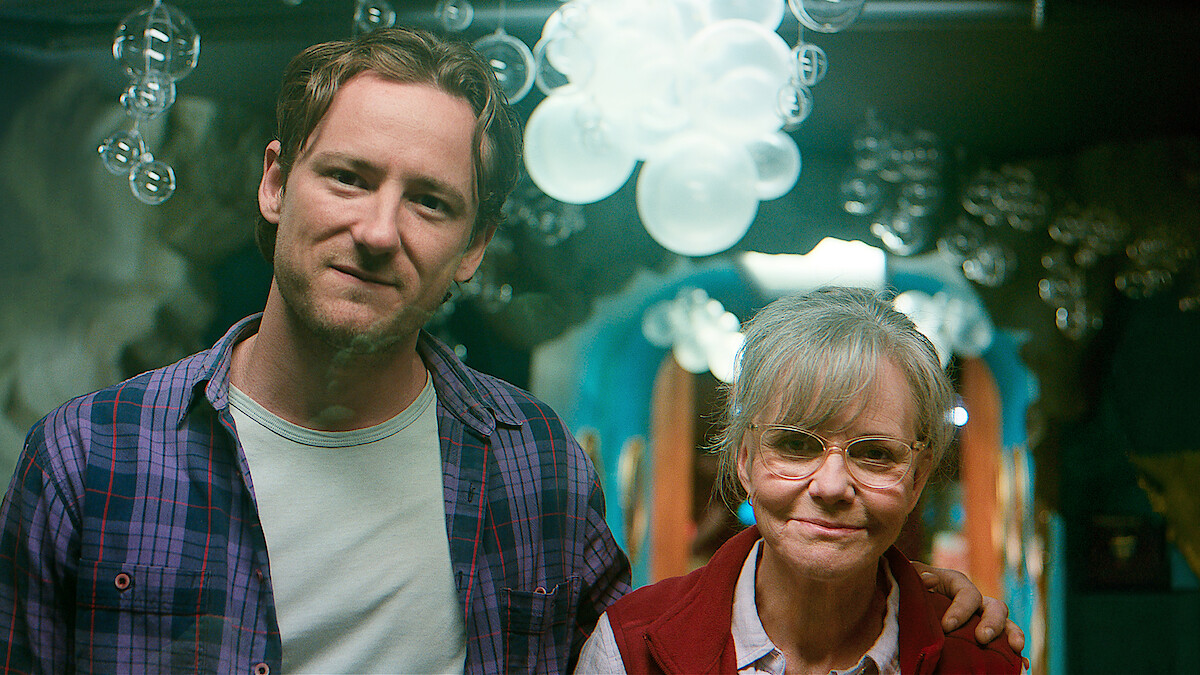 A younger man and an older woman stand together indoors, smiling at the camera, with his arm around her shoulders. The background features glass bubble decorations and soft, colorful lighting.