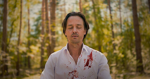 Tom Schilling sits in the woods meditating with his eyes closed, wearing a white shirt with blood splatter on it in an image from the series 'Murder Mindfully.'