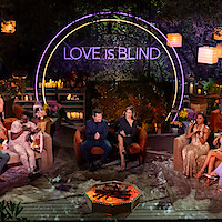 A group of well-dressed people sit in a semi-circle outdoors at night, under lanterns and trees, with a neon "Love Is Blind" sign behind them, creating a festive, lively, and glamorous TV show reunion atmosphere.