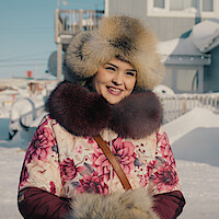 Woman smiling outdoors in snowy environment, wearing a floral coat and fur hat, with houses and snow-covered ground in the background, suggesting a cold winter setting.