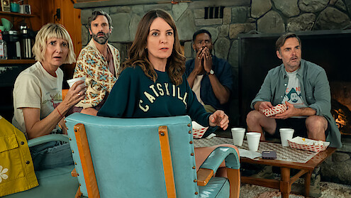 Five people sitting in a cozy living room with stone fireplace, snacks, and drinks, all looking surprised or shocked, suggesting an intense or suspenseful moment.