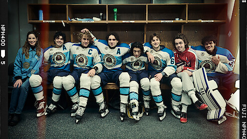 A group of young hockey players in blue and white uniforms and one in red sit together on a locker room bench, posing and smiling for a team photo, hockey gear and sticks by their sides.