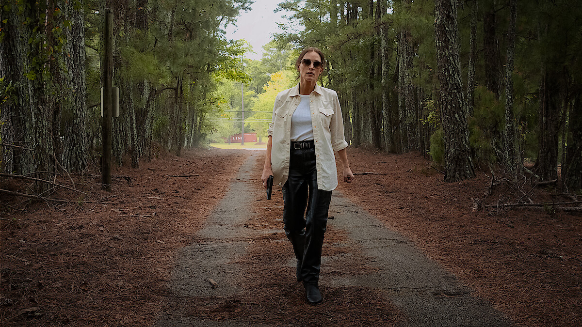 A woman in sunglasses and a white shirt, holding a gun, walks confidently down a forest path surrounded by tall trees, with sunlight filtering through the greenery in the background.