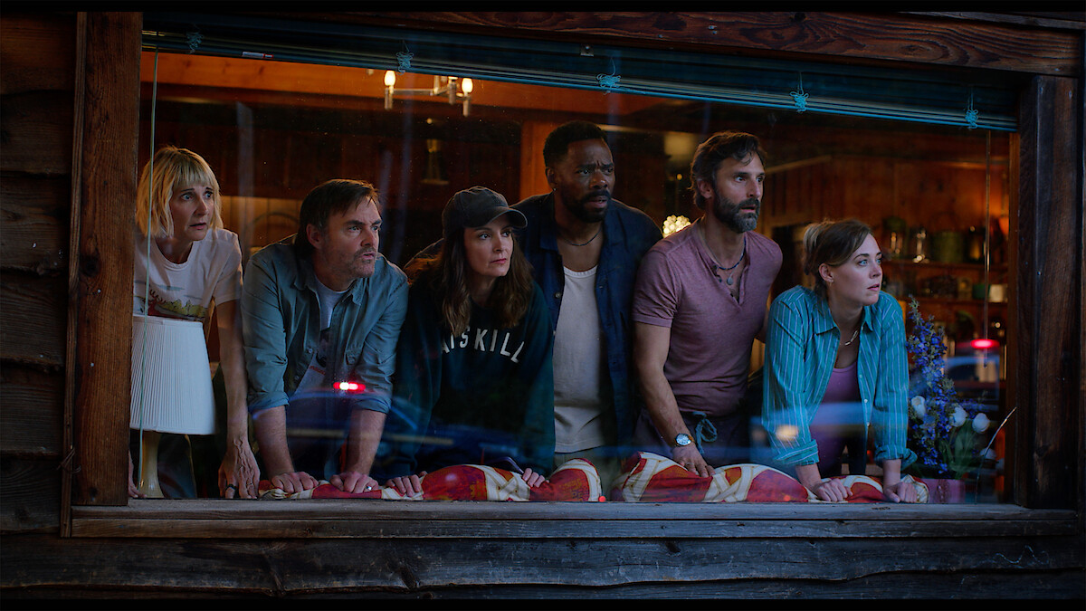 Six people lean forward together at a wooden cabin window, looking outside with concerned and curious expressions; the cozy, warmly lit interior contrasts with the dramatic mood.
