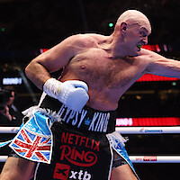 Tyson Fury in action mid-punch inside a brightly lit boxing ring, wearing gloves and trunks with "Gypsy King" and Union Jack flag, surrounded by an audience and stadium lights in the background.