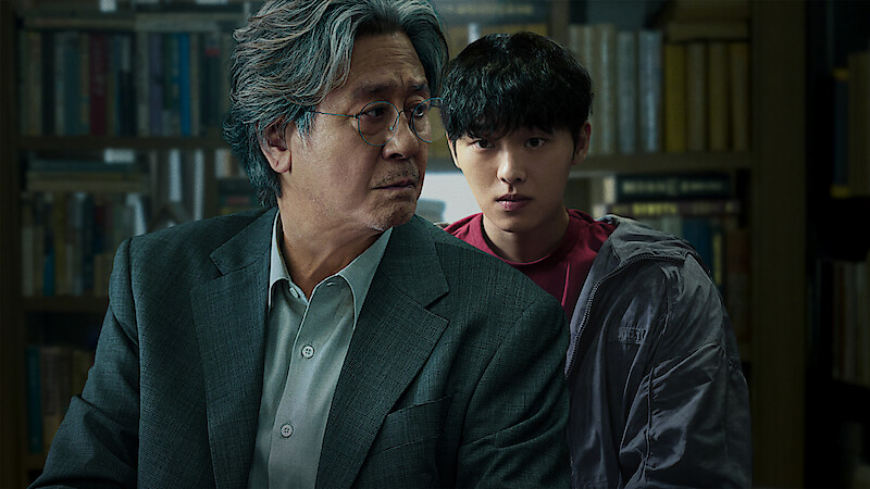 Two men, one older with gray hair in a suit and one younger with dark hair in casual clothes, are in a library or study filled with books, both appearing serious and focused.