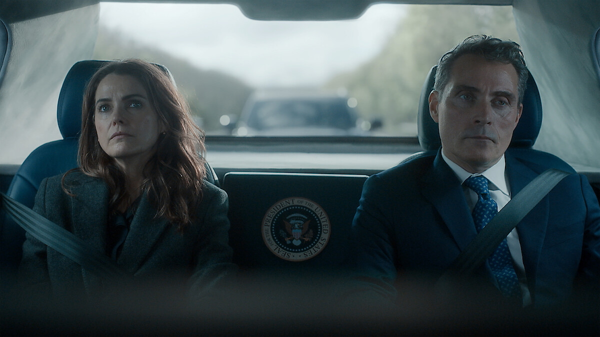 Two serious people, a woman and a man in formal attire, sit side by side in the backseat of a car with presidential seal, wearing seatbelts, with a blurred road and convoy vehicle visible through the rear window.