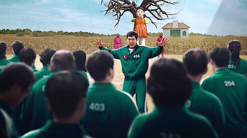 Group in green tracksuits faces man #456 with arms raised in a field. A giant doll, tree, and pink-clad guards are in the background near a small building.