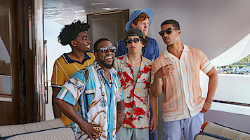 Five men in colorful summer shirts are standing and talking on a luxury boat deck, smiling and laughing together in a relaxed, sunny outdoor setting.
