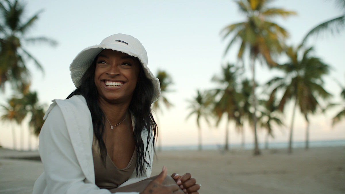 Simone Biles sorrindo enquanto está sentada na praia.