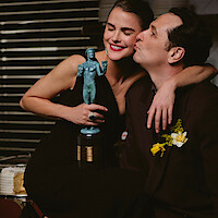 Keri Russell smiling and holding an award statuette while hugging Matthew Rhys, who is kissing her on the cheek, both dressed formally in an indoor setting with horizontal stripes in the background.
