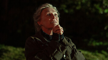 An older woman stands outdoors at night, hands clasped in front of her face as if in prayer, looking upward with a hopeful and contemplative expression, illuminated by warm light against a dark, blurred background.