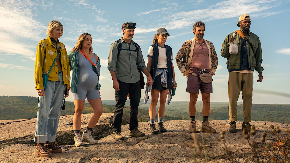 Six hikers stand on a rocky hilltop in casual outdoor gear, overlooking a scenic landscape under a blue sky with light clouds, suggesting a group hiking adventure in nature.