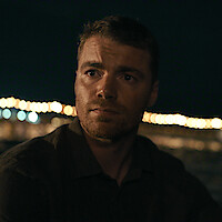 A man with short hair and a trimmed beard sits outdoors at night, illuminated by soft lights in the background, creating a pensive atmosphere with blurred string lights and dark sky.