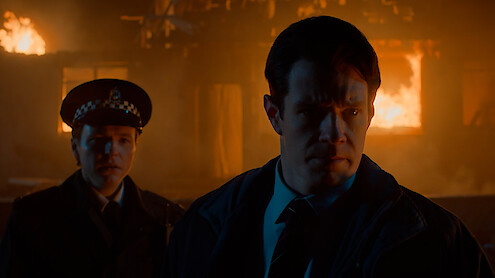 Two police officers stand outside at night with a burning building in the background, illuminated by firelight and smoke, appearing tense and serious as they respond to the emergency situation.