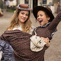 Two girls in vintage dresses and hats, one carrying the other while laughing, outdoors in what appears to be a historical or rural village setting with blurred buildings and people in the background.