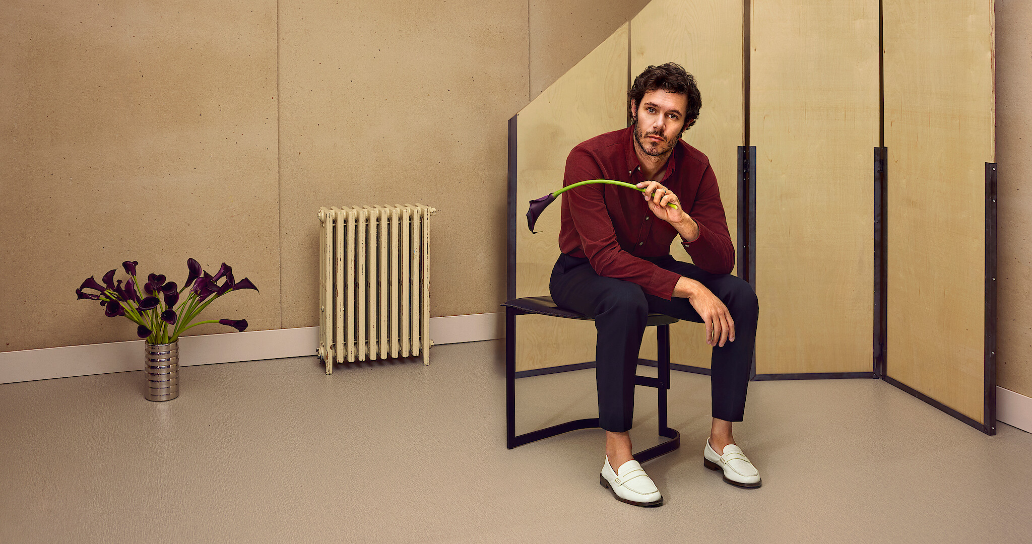 Adam Brody sits on a stool holding a black calla lily 