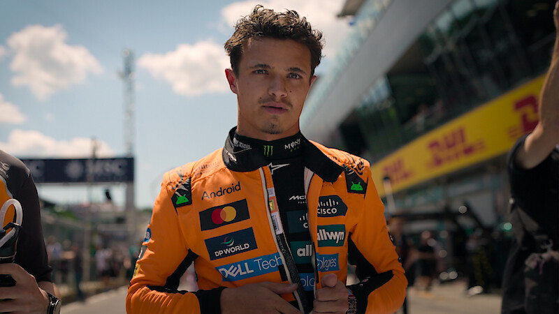 Lando Norris in an orange racing suit stands on a sunlit pit lane, surrounded by team members and blurred race track environment, preparing for a race.