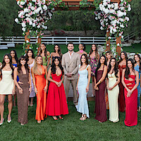A group of elegantly dressed women and one man stand together outdoors on grass, beneath a floral archway, with trees and a white fence in the background, posing for a formal photo.