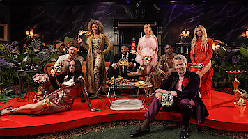 A group of elegantly dressed people holding masks pose in a lavish, dimly lit room with red carpets, gold decor, flowers, and lush plants, suggesting a masquerade party or formal event.