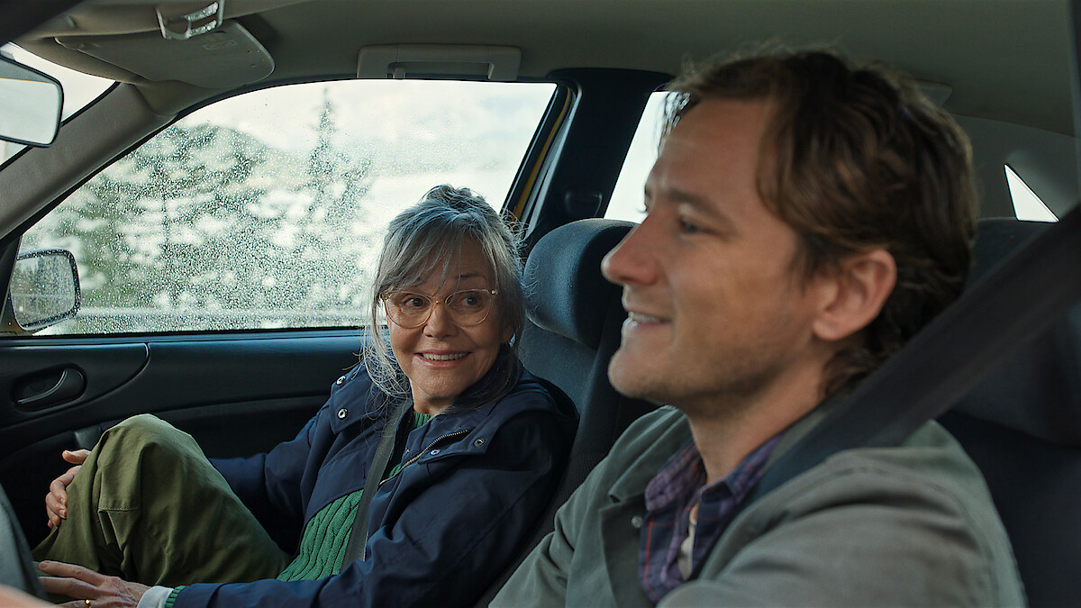 Two people sitting inside a car, one woman driving and smiling at the passenger, a man who is relaxed and smiling. The car is parked with a window showing a scenic outdoor background with trees and light rainfall.