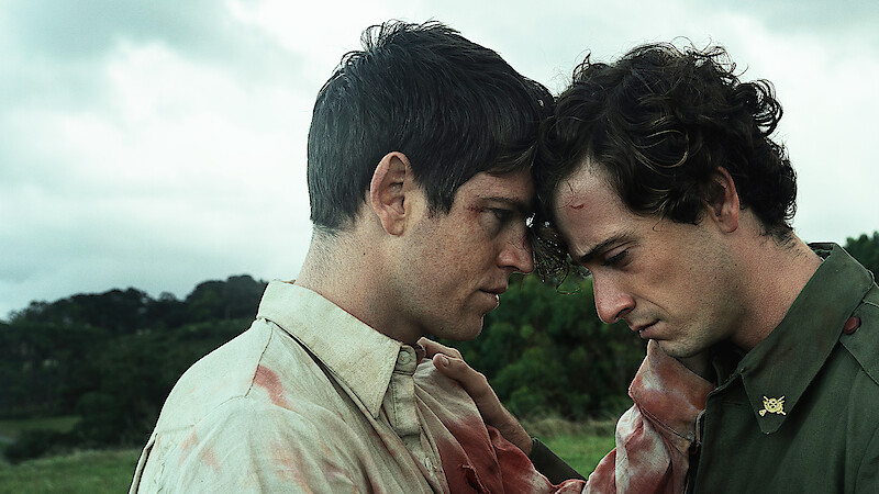 Two injured men with bloodied clothes stand close together in an emotional moment, foreheads touching, in an outdoor grassy field under a cloudy sky, suggesting a tense or dramatic scene.