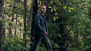 A man with gray hair and a beard walks through a dense, green forest wearing a dark jacket and jeans. The environment is lush with foliage, trees, and natural light filtering through the leaves.