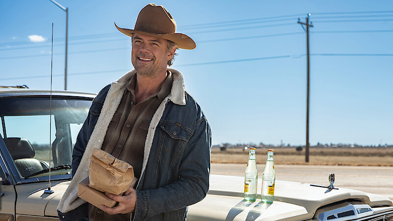 A man in a cowboy hat and denim jacket stands by a vintage truck, holding a paper bag. Two glass soda bottles rest on the truck, with power lines and an open rural landscape in the background.