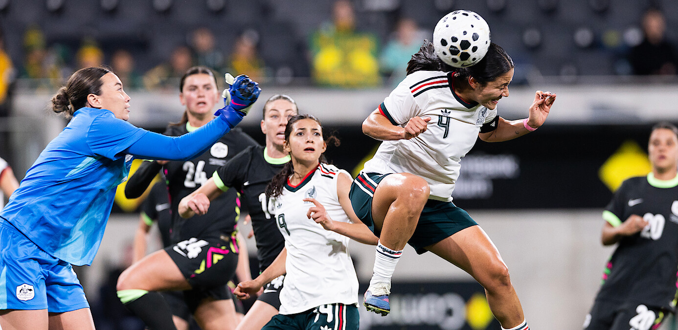 Women's soccer match in a stadium, goalkeeper in blue reaches for the ball as player in white and green jumps to head it, teammates and opponents in action, focused expressions, blurred spectators in background.