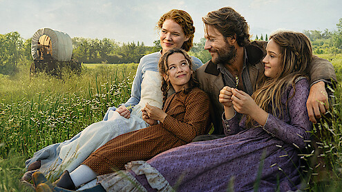 A family of four in historical clothing sits in a grassy field, smiling and enjoying time together, with a covered wagon and trees in the background on a sunny day.