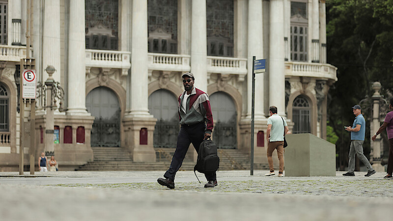 Man walking across a cobblestone plaza in front of a grand historic building with arched windows and columns, carrying a backpack; other people and street signs in the background.